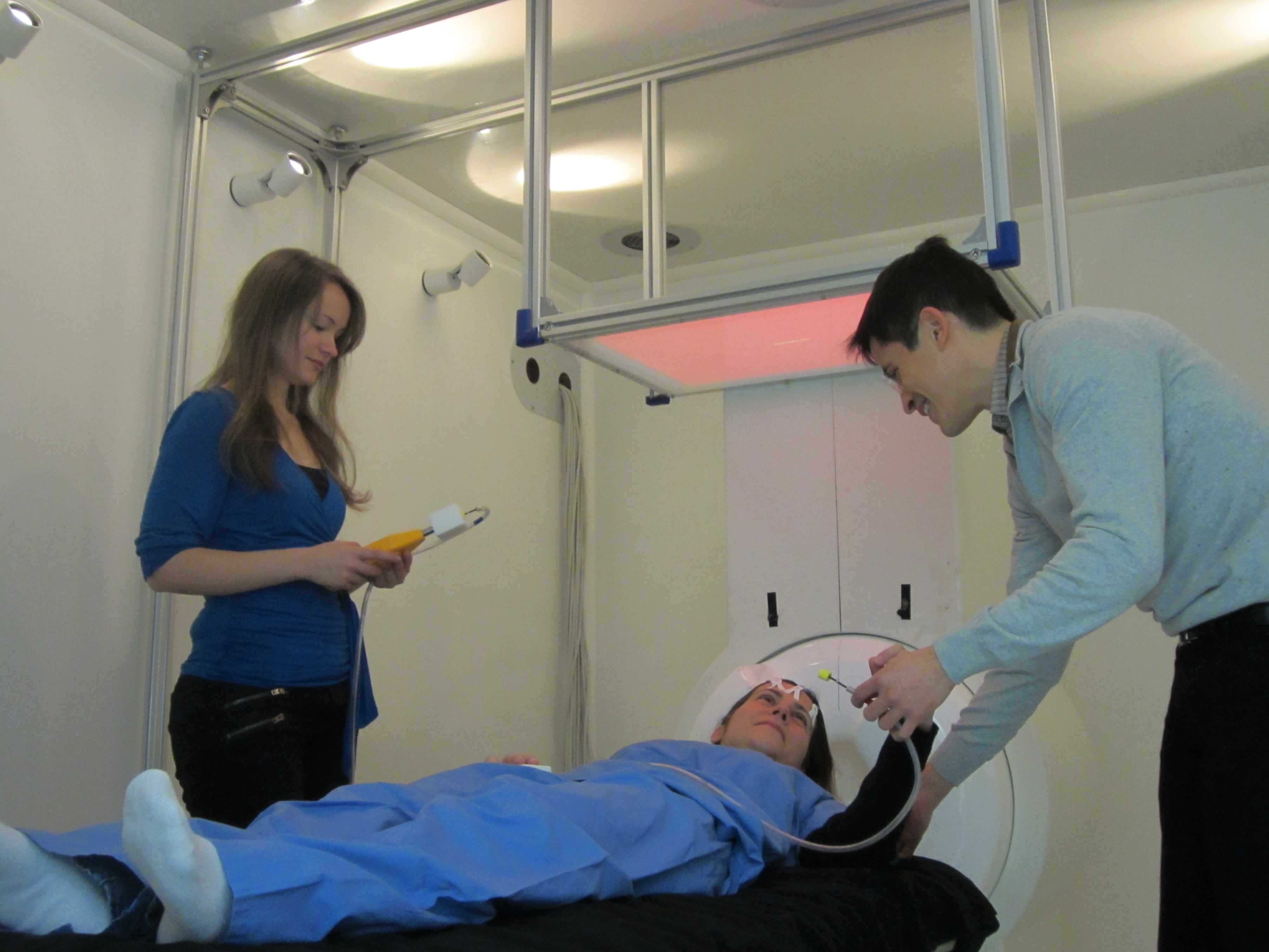 research participant laying down in an MEG scanner with two researchers attaching electrodes